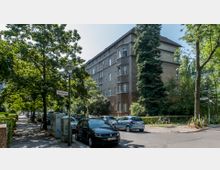 Wohnstraße in einer Stadt mit Bäumen entlang des Gehwegs, parkenden Autos und einem mehrstöckigen Wohngebäude aus grauem Putz mit roten Dächern im Hintergrund. Straßenschilder und eine Laterne sind sichtbar, umgeben von einer grünen, schattigen Umgebung.