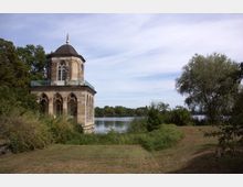 Pavillon im orientalischen Stil mit Rundkuppel und Spitzbogenfenstern, umgeben von Grünflächen und Bäumen, idyllisch an einem See gelegen. Im Hintergrund ist dichter Wald und ein ruhiger Himmel zu sehen.