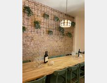 Ein rustikaler Raum mit einer Backsteinwand, an der Gitter befestigt sind, auf denen grüne Topfpflanzen hängen. Zentral steht ein langer Holztisch mit grünen Stühlen, darauf Weinflaschen, Gläser und dekorative Elemente; eine hängende Lampe mit rundem Schirm beleuchtet den Tisch.