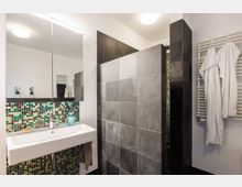 Modernes Badezimmer mit weißem Waschbecken und Spiegel, Mosaikfliesen in Grün- und Brauntönen an der Wand, grauen Fliesen in der Dusche und einem Handtuchheizkörper mit hängenden weißen Handtüchern.