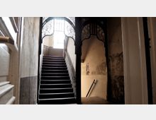 Historisches Treppenhaus mit abgenutzten Wänden, einem dunklen Holzgeländer und dekorativen, filigranen Metallverzierungen über der Treppe. Im Hintergrund ist ein großes Fenster, das Tageslicht hereinlässt.