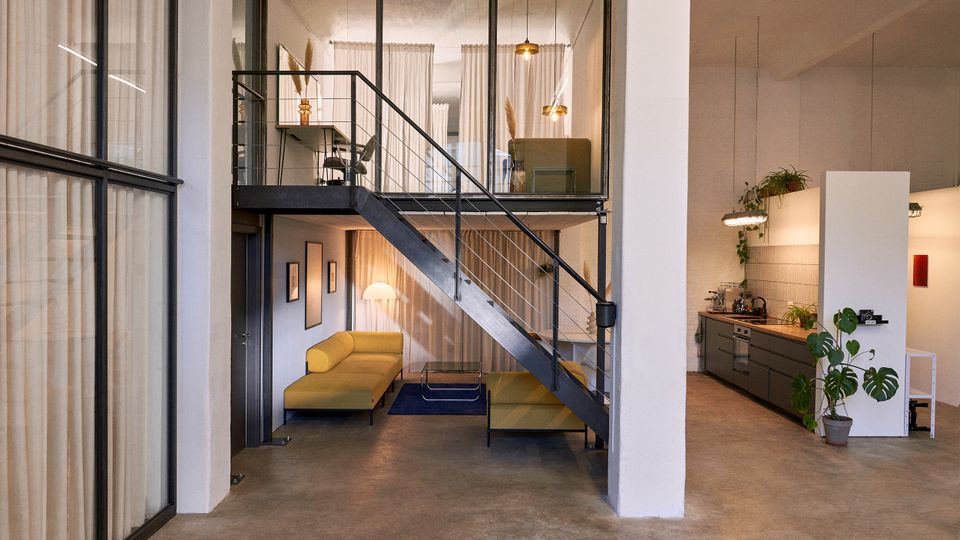 Offener Loft-Innenraum mit Betonboden, großer Glas-Trennwand und schwarzer Metalltreppe zu einer Galerieebene mit Vorhängen und Hängeleuchten. Unten befindet sich ein Sitzbereich mit Sofa, daneben eine moderne Küchenzeile mit Arbeitsplatte und Zimmerpflanzen.