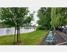 Uferbereich mit grünem Rasen, einigen kleinen Stroh-Sonnenschirmen und einem ruhigen Gewässer im Hintergrund. Auf einer Holzplattform rechts stehen bunte Liegestühle, daneben befinden sich Bäume, darunter eine große Trauerweide.