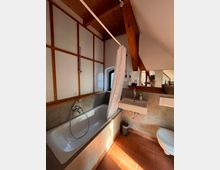 Badezimmer mit Dachschräge, ausgestattet mit weißer Badewanne, Duschvorhang, Wandwaschbecken und Toilette. Der Raum hat eine Kombination aus Holz- und Putzwänden, ein kleines Fenster sowie Terrakotta-Bodenfliesen.