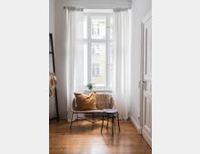 Sitzecke in einem hellen, schlichten Raum mit einem großen weißen Sprossenfenster, weißem Vorhang und Holzboden. Vor dem Fenster stehen ein filigraner Rattanstuhl mit Kissen und Decke sowie ein kleiner schwarzer Beistelltisch.