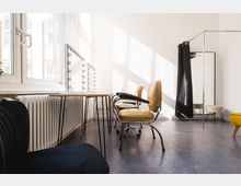 Heller Raum mit großem Fenster, in dem Tageslicht hereinfällt. Ein Schminktisch mit Spiegeln und runden Glühbirnen, mehrere Stühle mit beigen Polstern sowie ein schwarzer Vorhang und ein Ganzkörperspiegel prägen die Einrichtung.