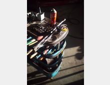 Werkstatt- oder Garagenbereich mit einem rollbaren Werkzeugwagen, auf dem Handwerkzeuge, Bitsätze, Schraubendreher und eine Spraydose liegen. Im Vordergrund ist eine Metallrolle mit Seil bzw. Kabel zu sehen, darunter mehrere Ablagefächer mit weiterem Werkzeug auf Betonboden.