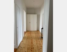 Ein heller Flur mit hellbraunem Holzboden und weiß gestrichenen Wänden. Im Hintergrund steht ein weißer Kleiderschrank, rechts daneben ein schmaler Beistelltisch mit schwarzem Metallgestell.