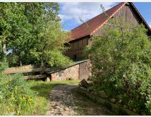 Ein ländliches Gebäude mit einer Fassade aus Holz und Ziegelsteinen sowie einem roten Ziegeldach, umgeben von dichten Büschen und Bäumen. Im Vordergrund führt ein gepflasterter Weg durch einen Garten mit Grasflächen und einem Holzzaun.
