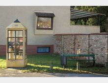 Gelbe Telefonzelle mit Graffiti vor einem Gebäude mit Putz- und Backsteinfassade, daneben eine grüne Parkbank und ein Mülleimer vor einer niedrigen Ziegelmauer. Im Hintergrund sind eine überdachte Terrasse und etwas Grün zu sehen.