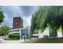 Backsteingebäude mit Uhrturm im Hintergrund und modernes, zweistöckiges Gebäude mit gläsernem Eingang im Vordergrund, umgeben von Bäumen, einer Grünfläche und Gehwegen. Der Himmel ist bewölkt, und eine Weide ragt ins Bild.