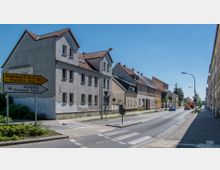 Straßenszene in einer Kleinstadt mit Wohnhäusern im traditionellen Baustil entlang einer zweispurigen Straße. Ein Verkehrsschild zeigt Richtungen nach Brandenburg an der Havel und Premnitz, während wenige Autos und ein Fußgängerüberweg sichtbar sind.