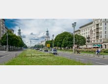 Weitläufige Straße mit Mittelstreifen aus Gras und Blumen, gesäumt von historischen Wohngebäuden mit großen Fenstern und Balkonen; im Hintergrund zwei Türme mit grünen Kuppeln und dem Fernsehturm von Berlin erkennbar. Entlang der Straße stehen Autos, Straßenlaternen und Bäume, Fußgänger und Radfahrer beleben die Szene.