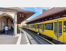 Ein U-Bahnhof mit gewölbten Dächern und gelben Zügen, die an einem Bahnsteig halten. Menschen stehen auf dem Bahnsteig, und im Hintergrund sind Gebäude und ein blauer Himmel sichtbar.