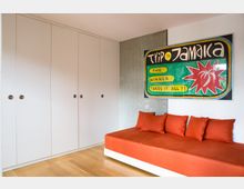 Ein schlichtes Zimmer mit weißem Einbauschrank, einer Wand aus Sichtbeton und einem roten Sofa mit dazupassenden Kissen. An der Wand hängt ein buntes Plakat mit der Aufschrift „Trip to Jamaica – The winner takes it all!!“.