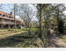 Großer Garten mit Bäumen und Sträuchern, angrenzend an ein zweistöckiges Wohnhaus mit rotem Schrägdach und Erkerfenstern; ein schmaler, von Bäumen gesäumter Pfad verläuft auf der rechten Seite.