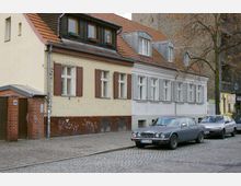 Straßenszene mit einer Reihe von Wohnhäusern mit unterschiedlichen Fassaden, darunter beige Kacheln, Holzläden und weiß gestrichene Elemente. Davor parkende Autos auf einer gepflasterten Straße, mit einem Baum am rechten Bildrand.