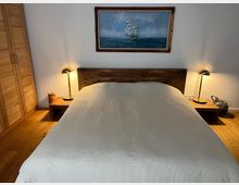 Schlafzimmer mit einem Holzbett in der Mitte, flankiert von zwei Nachttischen mit Lampen. Im Hintergrund hängt ein Gemälde mit einem Segelschiff, links steht ein Kleiderschrank aus Holz.