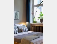 Ein modernes Schlafzimmer mit einem Doppelbett, das mit grauen und blaugestreiften Kissen sowie einer schlichten Tagesdecke dekoriert ist. Im Hintergrund befindet sich ein großes Fenster mit Vorhängen, ein Heizkörper darunter und Pflanzen sowie eine Vase auf der Fensterbank.