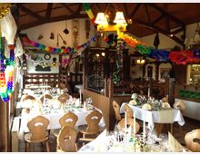 Innenraum eines rustikalen Restaurants mit Holzbalkendecke und dekorativen Vorrichtungen an den Wänden. Festlich gedeckte Tische mit weißen Tischdecken, Gläsern, gefalteten Servietten und buntem Partydekor, inklusive Girlanden und Luftschlangen, dominieren den Raum.