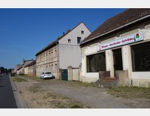 Straßenszene in einem ländlichen Ort mit einer Reihe von älteren Wohngebäuden aus hellem Mauerwerk und einem leerstehenden Geschäft mit dem Schild „Blumen-Geschenke-Getränke“. Die Straße ist asphaltiert, daneben verläuft ein sandiger Gehweg, und auf der rechten Seite steht ein geparktes Auto.