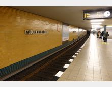 U-Bahn-Station Möckernbrücke in Berlin mit gelben Wandfliesen und einem Hinweisschild mit dem Stationsnamen. Rechts befinden sich Gleise, ein Bahnsteig mit wartenden Personen und eine Treppe, oben eine Anzeigetafel mit Linieninformationen.