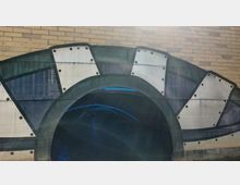 Ein gemalter Tunnelbogen mit geometrischen Mustern in Form von Dreiecken und Rechtecken auf einer Backsteinwand; im Inneren des Tunnels sind blaue Lichtstreifen sichtbar, die sich auf der Wasseroberfläche widerspiegeln.