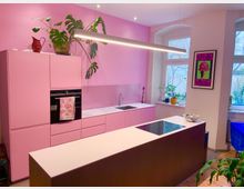 Moderne Küche mit rosa Wänden und rosa Schränken. Ein zentraler Küchenblock mit weißer Arbeitsfläche und eingebautem Kochfeld steht vor einer Wand mit Spüle, eingebautem Backofen und Pflanzen auf den Oberflächen. Ein großes Fenster und ein Poster schmücken den Raum.