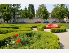 Ein gepflegter Park mit niedrigen, geometrisch geschnittenen Hecken, bunten Blumenbeeten und einem roten, modern gestalteten Sitzmöbel in der Mitte. Im Hintergrund stehen Bäume, eine beige Steinmauer und parkende Autos entlang einer Straße.