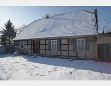 Vorderansicht eines eingeschossigen Fachwerkhauses mit großem, schneebedecktem Satteldach und kleinen Fenstern mit Fensterläden. Davor liegt ein verschneiter Hof, im Hintergrund stehen kahle Bäume und ein Zaun neben dem Gebäude.