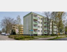 Mehrstöckiges Wohnhaus mit grün-weißer Fassade und Balkonen, umgeben von Bäumen und Rasenflächen. Im Vordergrund eine Straße, ein Gehweg sowie ein Tempo-30-Verkehrsschild.