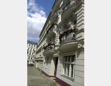 Wohnhaus im historischen Stil in einer engen städtischen Straße, mit verzierten Balkonen, Blumenarrangements und dekorativen Fassadenelementen. Im Hintergrund sind weitere ähnliche Gebäude und blauer Himmel zu sehen.
