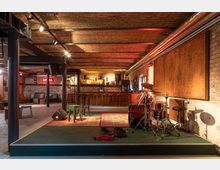 Industrie-inspirierter Veranstaltungsraum mit Ziegelwänden und freiliegenden Metallträgern, einer kleinen Bühne mit einem Schlagzeug und Kabeln sowie einer Bar im Hintergrund. Die Beleuchtung ist warm, und der Raum ist mit Teppichen und Holzmöbeln ausgestattet.