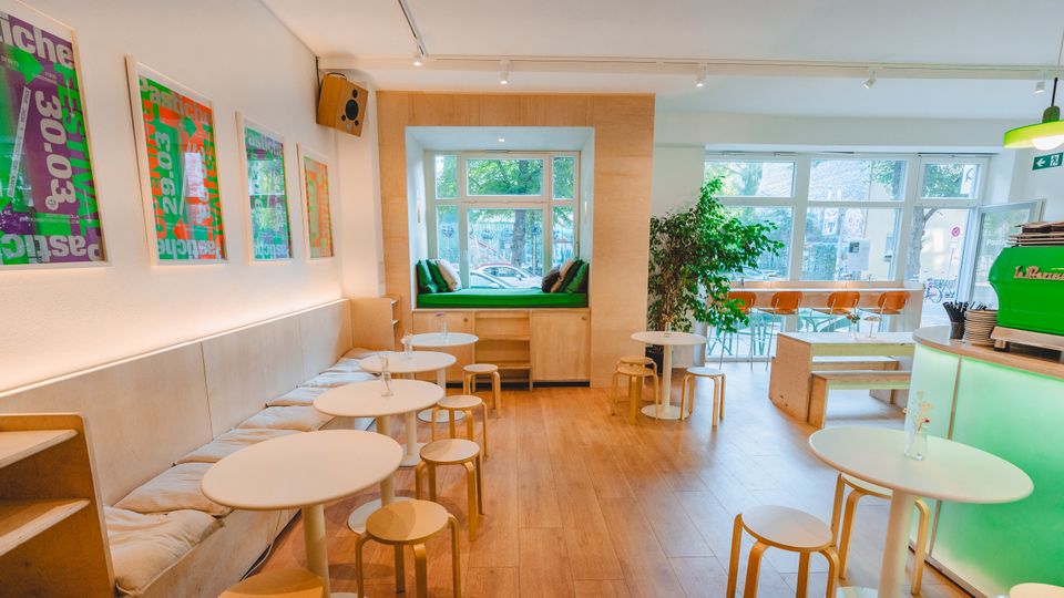 Bright, modern café interior with light wood floors, round white tables, and bench seating along a wall of framed posters. Large front windows include a built-in window seat with cushions, and a green espresso counter sits near the entrance with additional seating and indoor plants.