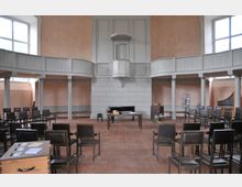 Innenraum einer schlichten Kirche mit cremefarbenen Wänden und weißen, umlaufenden Emporen auf Säulen. Der Raum ist mit Stühlen, einem schwarzen Flügel, einem Holztisch und einer Kanzel ausgestattet; seitlich ist eine Orgel sichtbar.