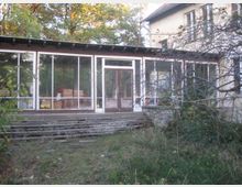 Ein verlassener Wintergarten mit großen Glasfronten und bröckelnder Treppe in bewachsener Umgebung, angrenzend an ein älteres Gebäude mit hellgelber Fassade. Im Inneren des Wintergartens sind Kartons und Gegenstände sichtbar.
