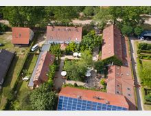 Luftaufnahme eines kleinen Komplexes aus roten Backsteinhäusern mit roten Ziegeldächern, teilweise mit Solarpaneelen bedeckt. In der Mitte befindet sich ein Innenhof mit Bäumen, Sonnenschirmen und Grünflächen, umgeben von ländlicher Umgebung und dichter Vegetation.