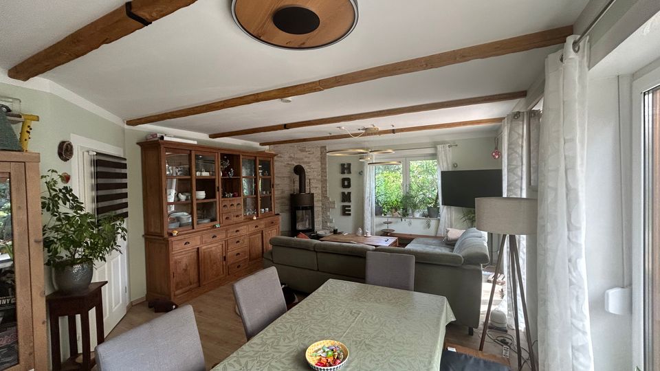 Offener Wohn- und Essbereich mit Holzbalkendecke, Esstisch im Vordergrund und angrenzender Sitzecke mit Ecksofa und Couchtisch. An der Rückwand stehen eine große Holzvitrine und ein Kaminofen, rechts sorgen breite Fensterfronten mit Vorhängen für viel Tageslicht.