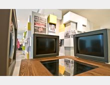 Zwei alte Computermonitore auf einem Holztisch in einem modernen Ausstellungssaal mit weißen und farbigen Wandmodulen sowie historischen Plakaten und Fotografien im Hintergrund.