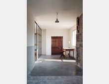 Ein minimalistischer Innenraum mit Betonboden, einer hölzernen Schiebetür an der Rückwand und einem schlichten Holztisch mit Hocker. Rechts ist eine unverputzte Ziegelwand, links eine Glaswand mit Metallrahmen, und von der Decke hängt eine einzelne Metalllampe herab.