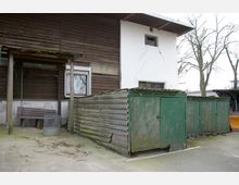 Außenbereich mit einem Gebäude aus Holz und verputztem Mauerwerk, daneben stehen mehrere grüne, verwitterte Wellblechschuppen. Im Vordergrund befindet sich eine überdachte Sitzbank und ein Müllkorb auf einem Betonboden.