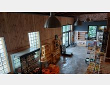 Ein moderner Hofladen mit einer Holzwandverkleidung, freiliegenden Ziegeln und großen Metallpendelleuchten. Das Innere zeigt Regale mit Lebensmitteln, Getränkekisten und Produkten sowie eine Glastür, die ins Freie führt.