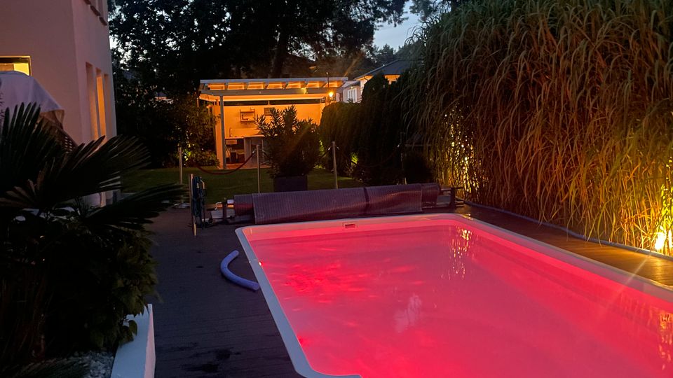 Ein beleuchteter Außenpool mit rotem Licht in einer Gartenanlage, umgeben von hohen Gräsern und Bäumen, mit einem modernen Gebäude und einer Terrasse im Hintergrund.