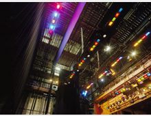 Blick hinter die Bühne eines Theaters mit einem hohen Raum, Gitterstrukturen an der Decke und zahlreichen farbigen Scheinwerfern an riggenartigen Aufhängungen. Im Vordergrund sind Bühnentechnik und Vorhänge zu sehen.