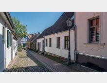 Gepflasterte Gasse in einer Wohngegend mit historischen Gebäuden, die durch Fachwerk- und Putzfassaden geprägt sind; ein Laternenmast und ein Baum im Hintergrund ergänzen die Szenerie.