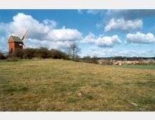 Eine hügelige Wiese mit einer roten Windmühle mit weißen Flügeln auf der linken Seite, umgeben von Büschen und Bäumen unter einem teils bewölkten Himmel. Im Hintergrund ist eine kleine Siedlung mit Häusern und Feldern zu sehen.