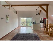 Heller Wohnraum mit offener Küche, sichtbaren Holzbalken und großen Glastüren, die auf einen Balkon mit Blick ins Grüne führen. Der Raum hat Fliesenboden, eine Kochinsel und eine Holztreppe auf der rechten Seite.