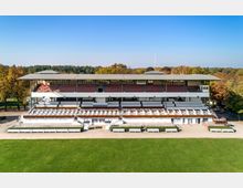 Zweistöckige Tribüne einer Pferderennbahn mit rot-weißen Sitzreihen, überdachtem Bereich und Blumenschmuck an den Balustraden. Im Vordergrund befinden sich gepflegte Grünflächen, umgeben von herbstlich gefärbten Bäumen im Hintergrund.