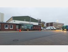 Straßenansicht eines Gewerbegebäudes mit einer Backsteinfassade und einem Restaurant im Vordergrund. Im Hintergrund sind eine Halle mit grün-weißer Außenwand und mehrere geparkte Fahrzeuge sowie ein vorbeifahrender Lastwagen zu sehen.