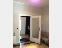 Heller Flur mit offen stehender Zimmertür aus Holz und Glaseinsatz, grauen Wänden und beigefarbenem Bodenfliesen mit schwarzem Randmuster. Rechts stehen zwei schlichte gepolsterte Stühle, links ist ein Heizkörper und eine Duftstäbchenhalterung zu sehen.